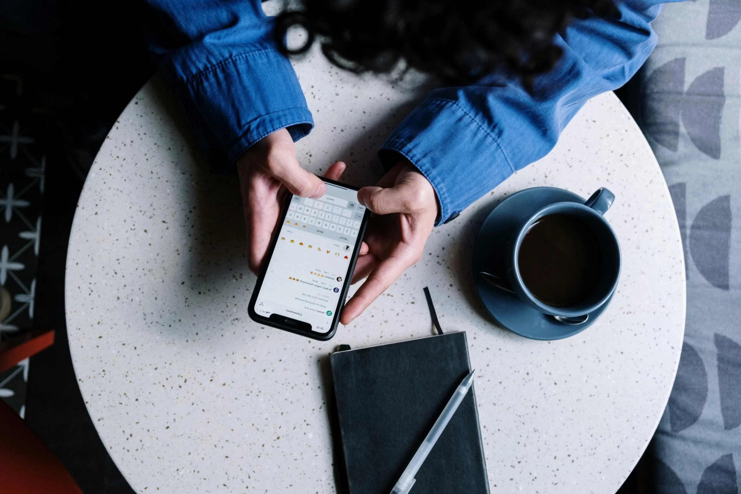 Photo of person using a phone while with a cup of coffee and notebook in a table