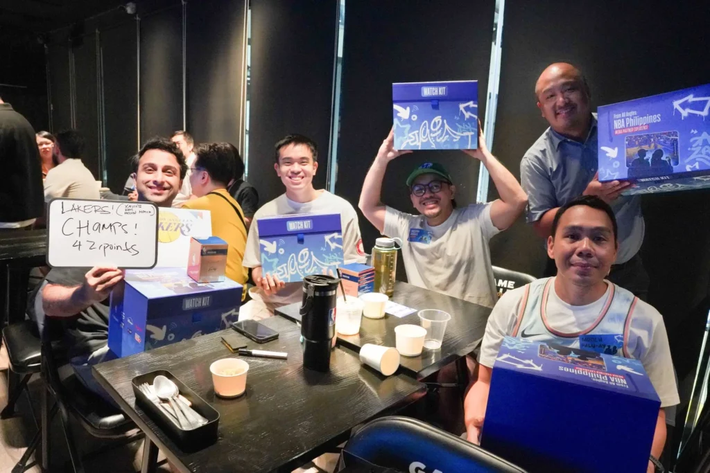 A group of NBA Philippines trivia winners proudly display their Watch Kit prizes at Gameville Sports Bar, as documented in M2.0 Communications' PR campaign case study. The whiteboard reads "Lakers! Champs! 42 points.