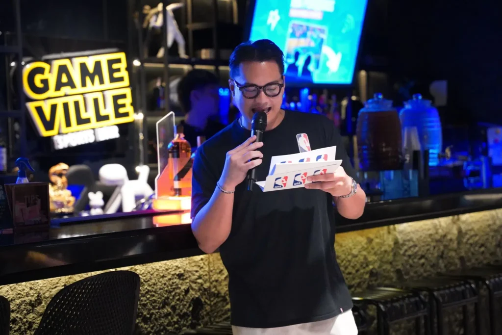 A host holding an NBA-branded envelope speaks into a microphone at Gameville Sports Bar during an NBA Philippines fan event, featured in M2.0 Communications' PR campaign case study.
