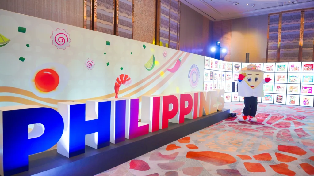 The Lucky Me! mascot stands beside large 3D "Philippines" lettering at a "World United by Noodles" event backdrop, as documented in M2.0 Communications' PR campaign case study.