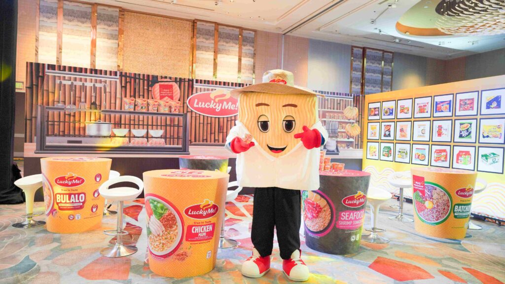 The Lucky Me! noodle mascot poses among oversized product displays of Bulalo, Chicken Mami, Seafood Shrimp Tonkotsu, and Batchoy flavors at a brand launch event, featured in M2.0 Communications' PR campaign case study.